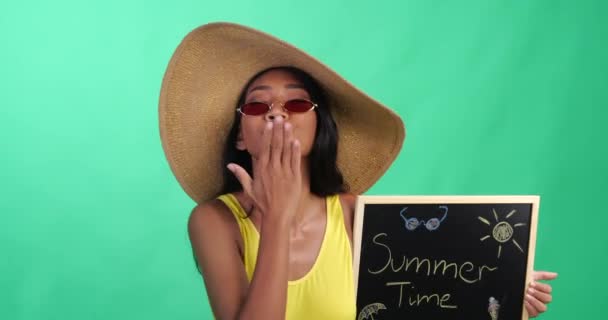 Heldere zomer portret van mooie lachende vrouw in gele badpak, hoed en rode zonnebril Holding Chalk Board met zomertijd schrijven geïsoleerd over groen schermachtergrond - Video