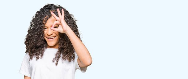 Young beautiful woman with curly hair wearing white t-shirt doing ok gesture with hand smiling, eye looking through fingers with happy face. - Foto, Bild