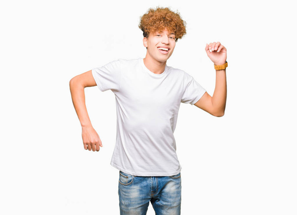 Jóképű fiatalember visel alkalmi fehér póló tánc, boldog és vidám, mosolygós, mozgó, alkalmi és magabiztos hallgató-hoz zene afro haj - Fotó, kép