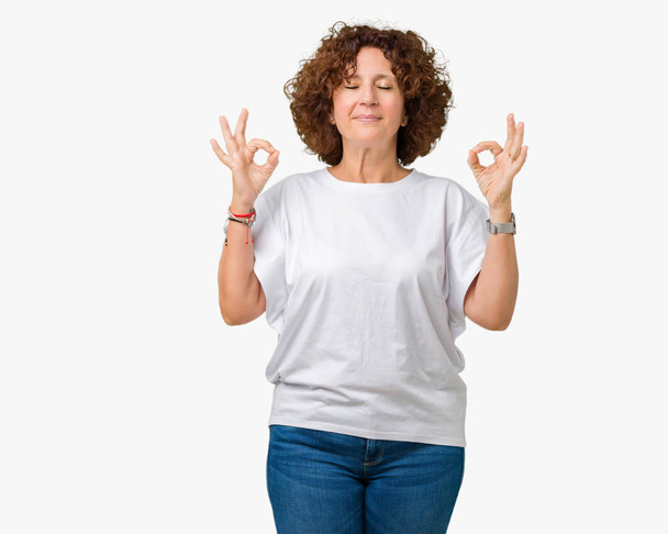 Beautiful middle ager senior woman wearing white t-shirt over isolated background relax and smiling with eyes closed doing meditation gesture with fingers. Yoga concept. - Foto, afbeelding