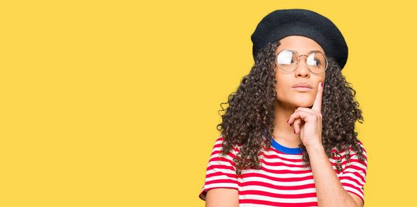 Young beautiful woman with curly hair wearing glasses and fashion beret with hand on chin thinking about question, pensive expression. Smiling with thoughtful face. Doubt concept. - 写真・画像