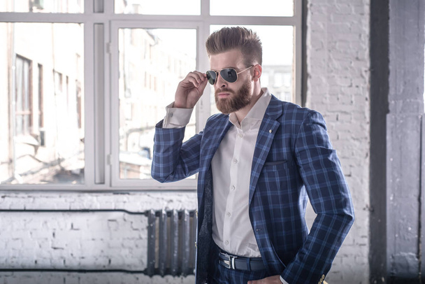 Time to go. Portrait of handsome young man in full suit and suspenders looking away and keeping sunglasses by his hand while standing in the loft interior - Foto, imagen
