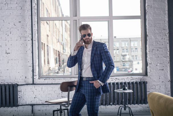 Looking perfect. Full length of handsome young bearded man in full suit and sunglasses looking to the camera while speaking by his cell phone. standing at loft interior - Photo, Image