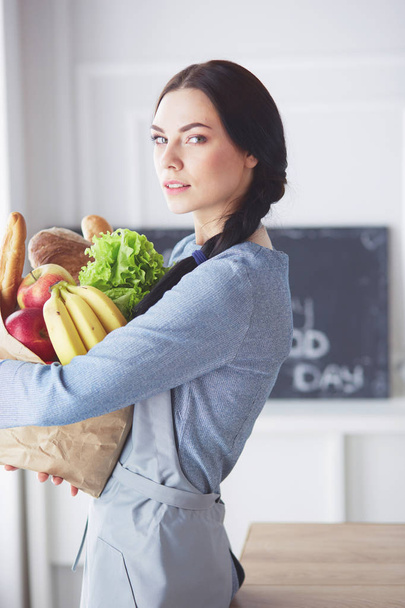 Young woman holding grocery shopping bag with vegetables .Standing in the kitchen. - Фото, изображение
