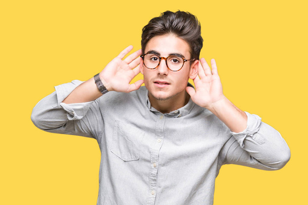 Молодой красивый мужчина в очках на изолированном фоне пытается услышать жесты обеих рук, любопытные для сплетен. Проблемы со слухом, глухой
 - Фото, изображение