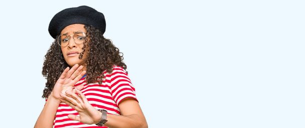 Young beautiful woman with curly hair wearing glasses and fashion beret disgusted expression, displeased and fearful doing disgust face because aversion reaction. With hands raised. Annoying concept. - Foto, imagen