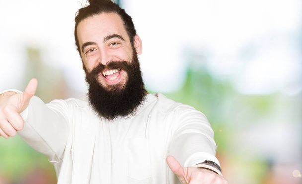 Young man with long hair and beard wearing sporty sweatshirt approving doing positive gesture with hand, thumbs up smiling and happy for success. Looking at the camera, winner gesture. - Φωτογραφία, εικόνα