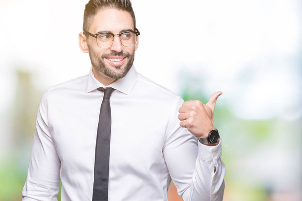 Young handsome business man wearing glasses over isolated background smiling with happy face looking and pointing to the side with thumb up. - Foto, Imagen