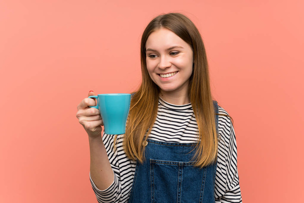 junge Frau mit Overalls über rosa Wand hält eine heiße Tasse Kaffee - Foto, Bild