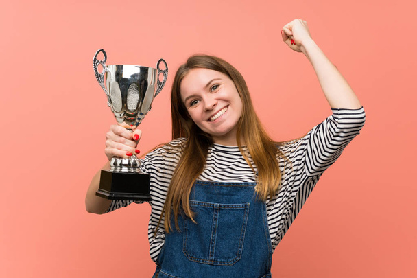 Young woman with overalls over pink wall holding a trophy - Foto, Bild