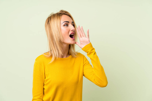 Jonge blonde vrouw over geïsoleerde groene achtergrond schreeuwen met mond wijd open - Foto, afbeelding