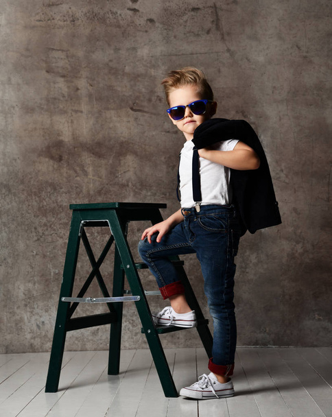 Cute stylish boy sitting on chair near concrete wall - 写真・画像
