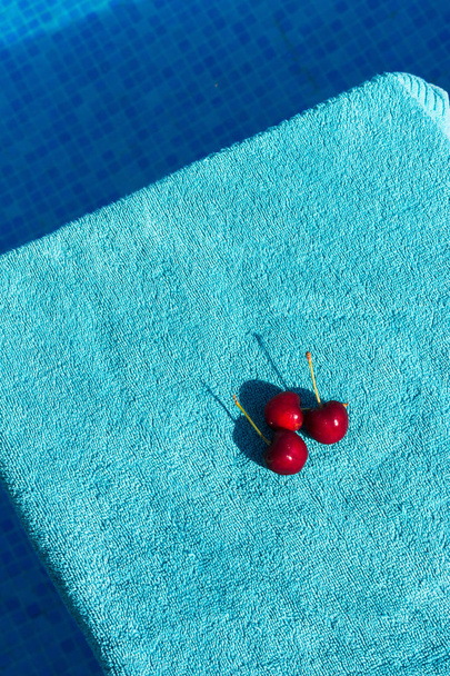Tres cerezas rojas profundas en una superficie azul texturizada justo al lado de una piscina
 - Foto, imagen