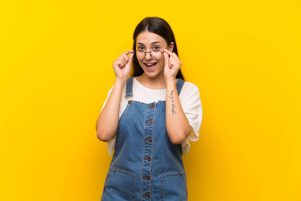 Young woman in dungarees over isolated yellow background with glasses and surprised - Photo, Image