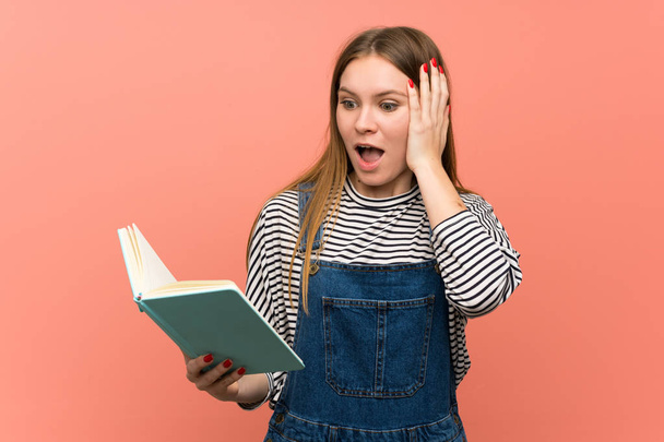 Young woman with overalls over pink wall surprised while enjoying reading a book - Fotoğraf, Görsel