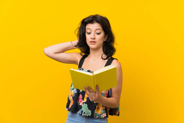 Young woman over isolated yellow background holding and reading a book - Фото, изображение