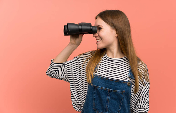 Young woman with overalls over pink wall and looking in the distance with binoculars - Foto, Bild