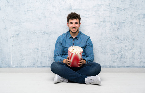 Nuori mies istuu lattialla popcorneja kädessään.
 - Valokuva, kuva