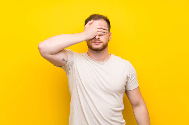 Handsome man over yellow background covering eyes by hands - Foto, imagen