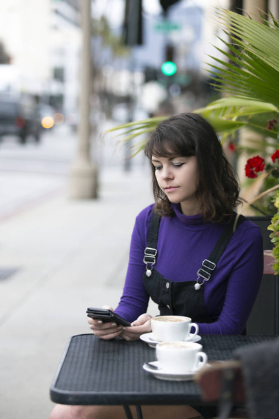 コーヒーショップやレストランの外に座っている女性は、待っている間、またはたむろしている間、携帯電話でソーシャルメディアを閲覧しています。彼女はまた、カフェの食べ物やサービスを評価したり、レビューすることができます.  - 写真・画像