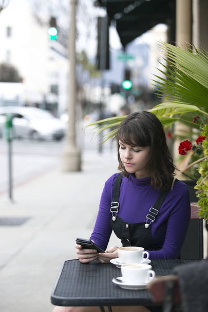 コーヒーショップやレストランの外に座っている女性は、待っている間、またはたむろしている間、携帯電話でソーシャルメディアを閲覧しています。彼女はまた、カフェの食べ物やサービスを評価したり、レビューすることができます.  - 写真・画像