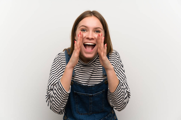 Young woman in dungarees over white wall shouting with mouth wide open - Photo, Image
