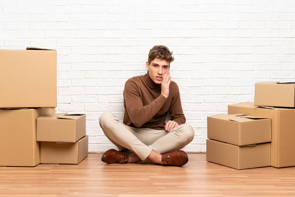Όμορφος νεαρός άντρας που μετακομίζει σε νέο σπίτι ανάμεσα σε κουτιά που ψιθυρίζουν κάτι - Φωτογραφία, εικόνα