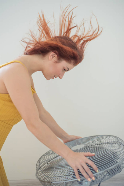 Portrait of a beautiful red-haired woman in a mustard jumpsuit enjoying the cooling breeze from a large electric fan. A smiling girl freshens up in the hot summer heat. Hair develops from the wind - Foto, Bild