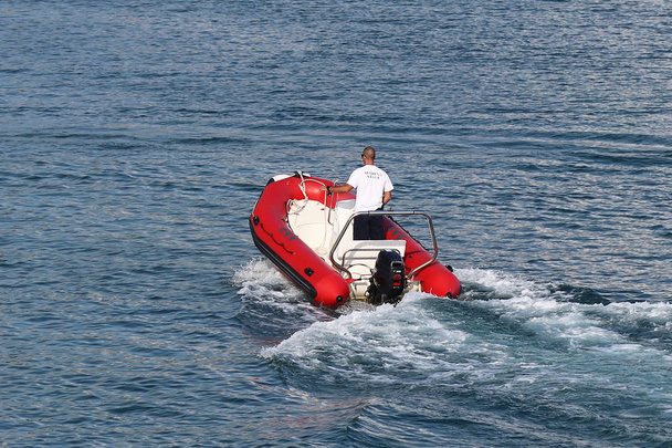 O staf marina está navegando em um barco a motor inflável vermelho para um iate de cruzeiro à vela que entra na marina. Assistência de pilotagem com a amarração de um barco fretado. Verificação do navio pela guarda
 - Foto, Imagem
