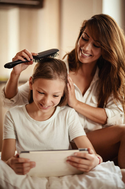 Young smiling happy mother combing little daughter hair on bed  - Foto, immagini
