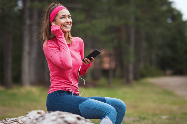 Young female runner listening to music and taking break after jogging in sunny forest - Фото, изображение