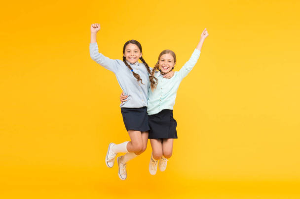 Happy winners. Little school girls jumping and making winner gestures on yellow background. Cute small winners enjoying happiness. Energetic winners celebrating victory or success togethers - Фото, изображение