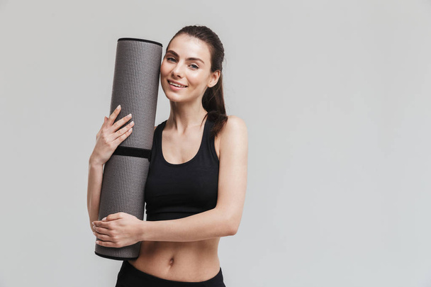 Beautiful young sport fitness woman make exercises isolated over grey wall background holding fitness rug. - Foto, immagini