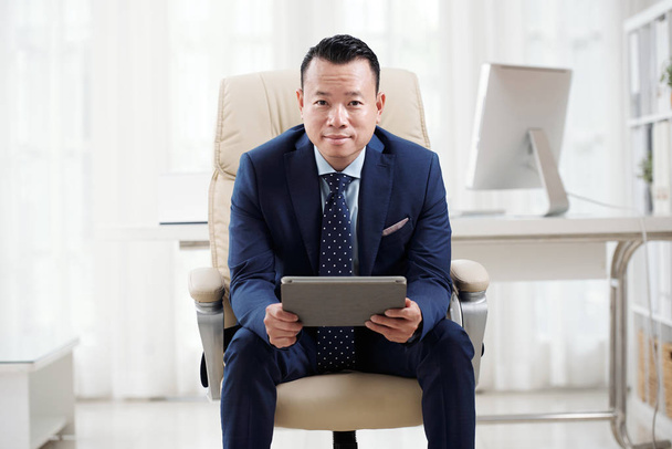 Porträt eines selbstbewussten asiatischen Geschäftsmannes, der auf einem Stuhl sitzt, ein digitales Tablet in der Hand hält und im Büro in die Kamera schaut. - Foto, Bild