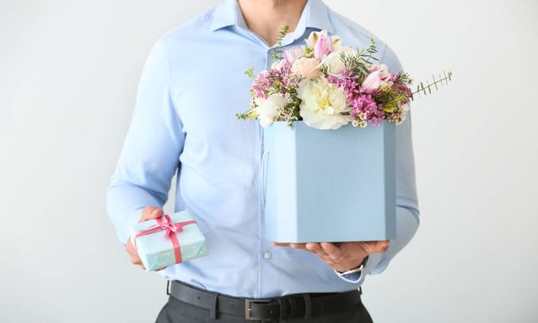 Handsome man with bouquet of beautiful flowers and gift box on light background - Photo, Image