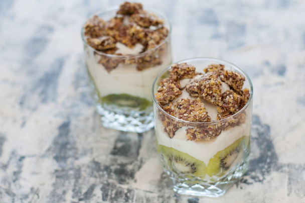 Granola, kiwi, banana e iogurte grego em duas xícaras de vidro em concreto cinza, espaço de cópia. Fitness, figura, corpo e comida saudável
 - Foto, Imagem