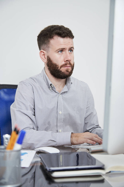 Серьезный бородатый мужчина сидит за столом и смотрит на монитор компьютера и печатает на клавиатуре компьютера в офисе
 - Фото, изображение