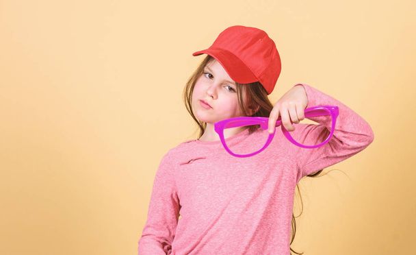 Stylish accessory. Feeling confident with accessories. Girl cute child wear cap or snapback hat hold eyeglasses beige background. Little girl wearing bright baseball cap. Hat or cap. Modern fashion - Фото, изображение