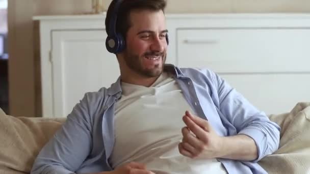 Happy man sitting on couch wearing headphones listening to music - Záběry, video