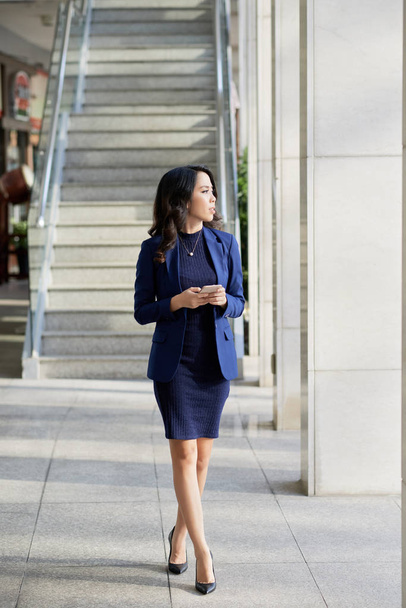 Asiática joven empresaria en ropa formal sosteniendo el teléfono móvil y mirando hacia otro lado mientras camina en la ciudad
 - Foto, imagen