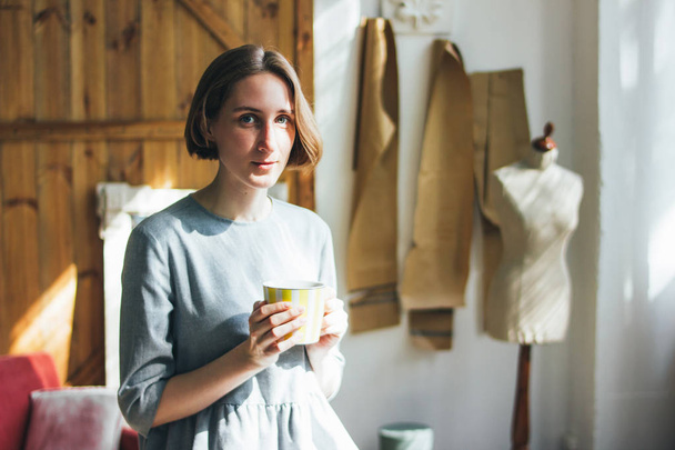 Young woman in simple grey dress with yellow cup sitting on chair in art work studio, full length portrait - Foto, Imagem
