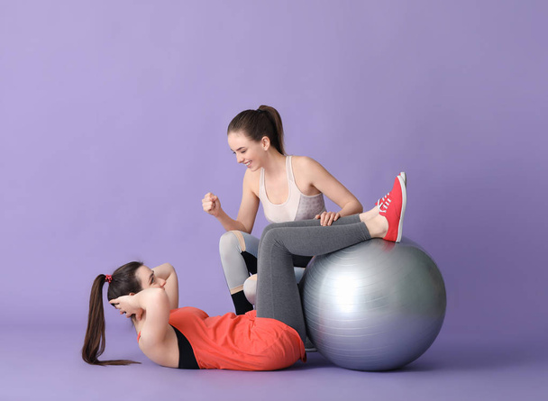 Young sporty woman with fitball doing exercises under supervision of her personal trainer on color background - Foto, Imagen