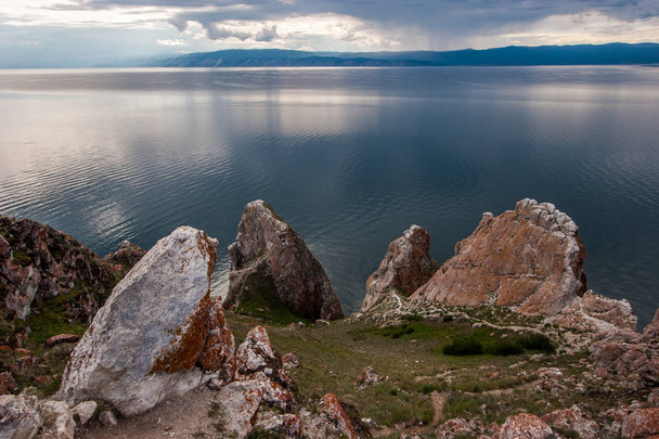 Piękne duże kamienie na tle jeziora Bajkał. Na kamieniach jest czerwony mech. W chmurach nieba. Za jeziorem są góry i deszcz. - Zdjęcie, obraz