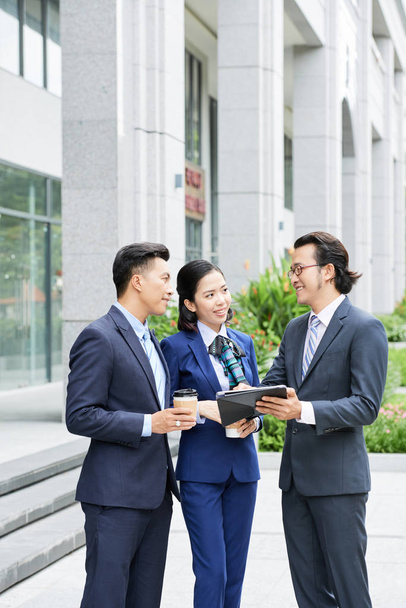 Homme d'affaires asiatique en costume et lunettes debout avec tablette numérique et discuter avec ses partenaires projet en ligne dans la ville en plein air
 - Photo, image