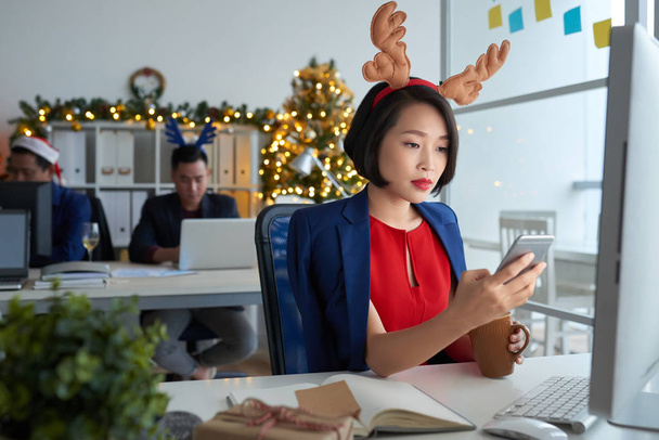 Pensive concentrated young Asian lady in antler headbands sitting at table in front of computer and texting sms while working in office and drinking coffee on eve of New Year - Photo, Image