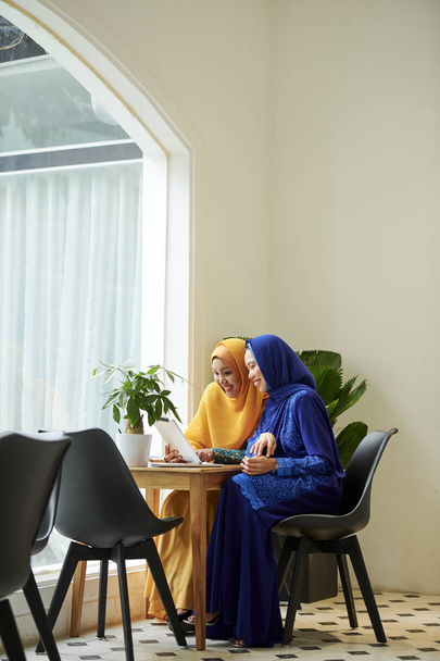 Θετικές νεαρές μουσουλμανικές γυναίκες σε φωτεινά πολύχρωμα φορέματα που κάθονται στο καφέ τραπέζι και Shopping online μέσω υπολογιστή tablet - Φωτογραφία, εικόνα