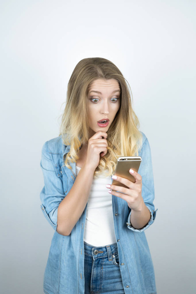 blond young woman looks astonished at smartphone in her hand isolated white background - Foto, afbeelding