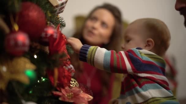 Mother, father and little baby sitting near decorated christmas tree close up. Man holding child near fir tree, showing bright decoration. Happy family celebrates Christmas together. Slow motion. - Video, Çekim