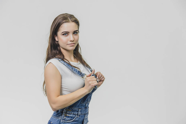 A cheerful cute teen girl holds his hands on the pliers. Has a surprised facial expression and looks into the camera. Dressed in white t-shirt and denim overalls. - Photo, image