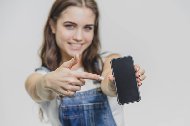 Young pretty girl standing on a white background. Shocked beautiful girl in denim overalls. Looking at the camera in amazement and admiration. Holding the phone on the girls blurry background - Фото, зображення